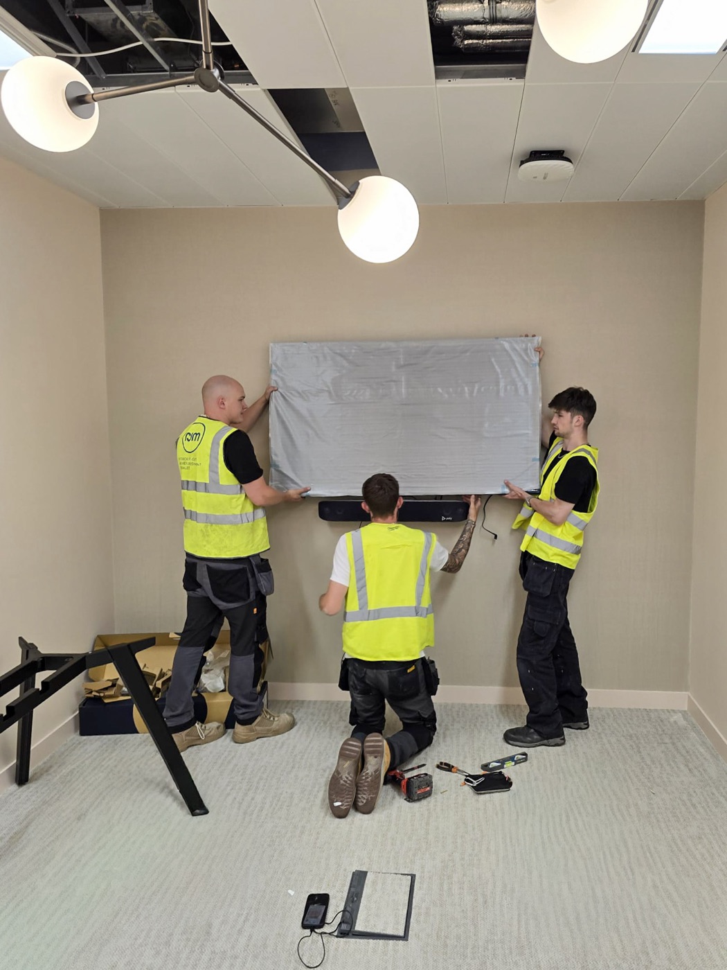 Three MAV engineers in hi-vis lifting a protected display onto its wall mount above a Poly video bar