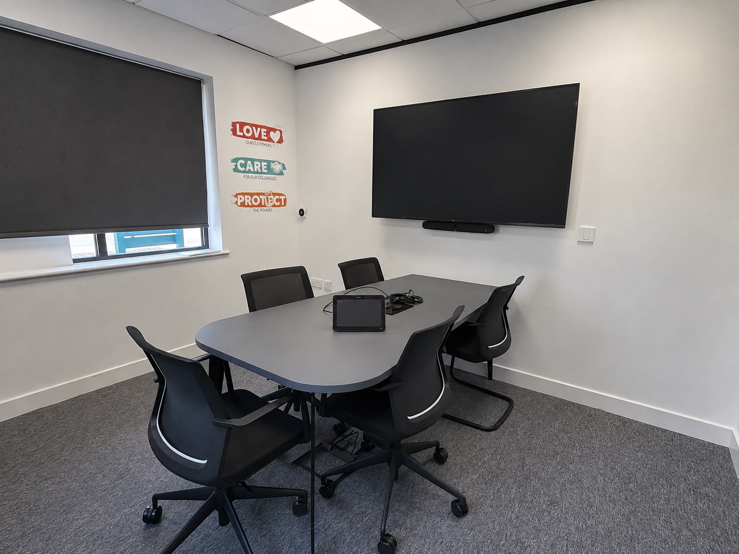 Meeting room with a wall-mounted display and video bar, touch controller on the table and brand values wall graphics